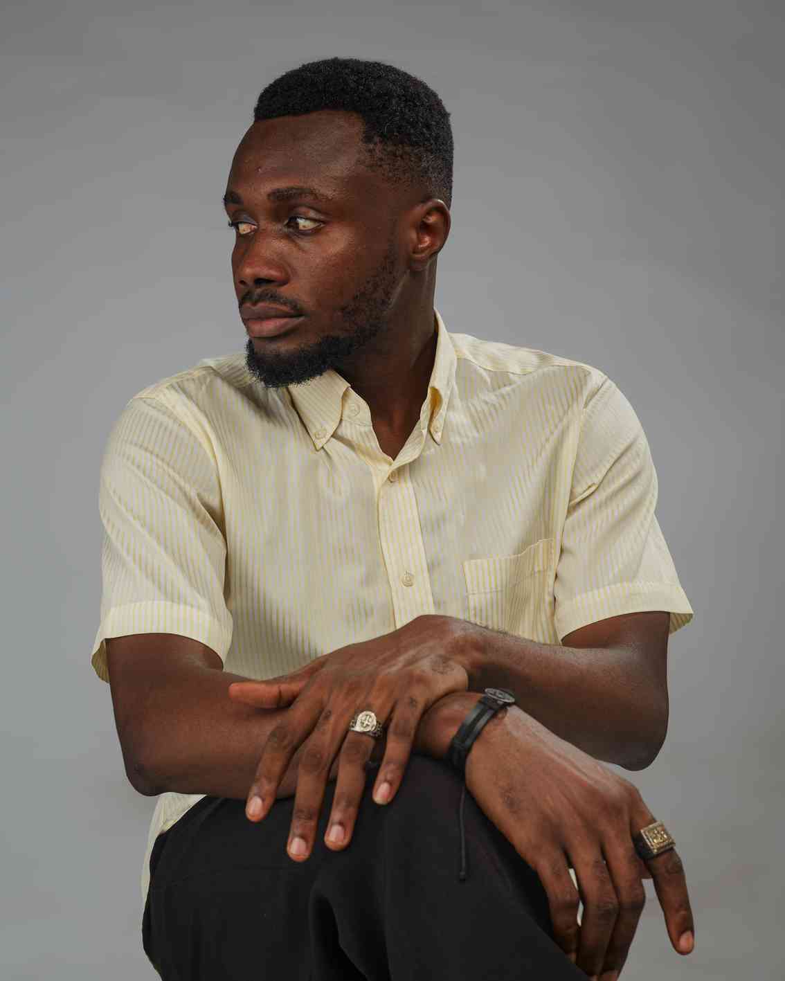 A man in a light striped shirt seated with his arms crossed, looking away from the camera. His pose is relaxed yet alert, and his facial expression suggests attentiveness or reflection.