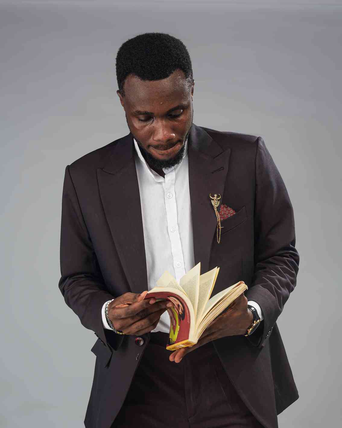 A well-dressed man in a dark suit and white shirt reads a book while standing. The angle and his downcast gaze convey a sense of quiet concentration and intellect.