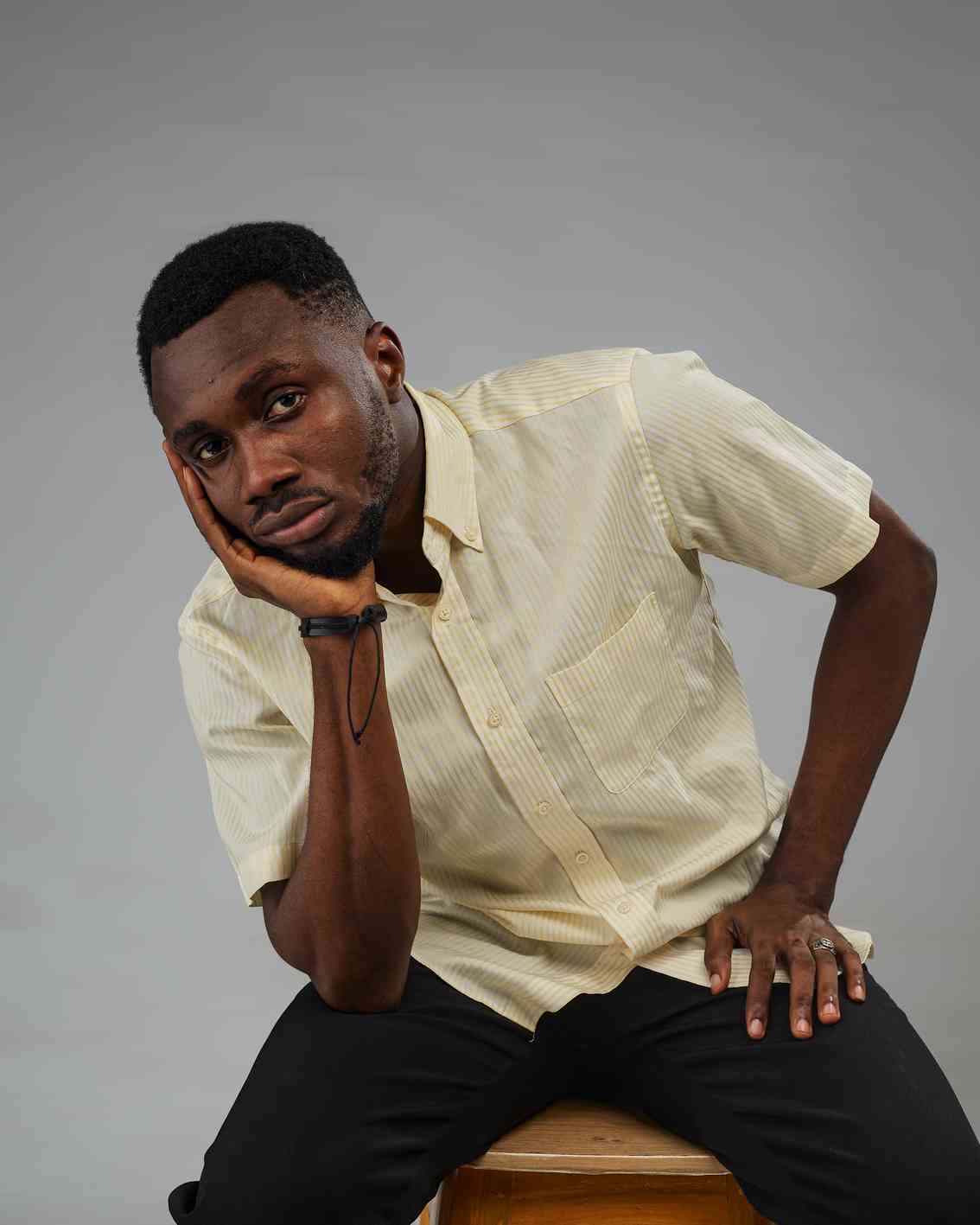 A young man in a black t-shirt shrugs his shoulders with palms up, looking upwards in a confused or uncertain expression. Neutral grey background.