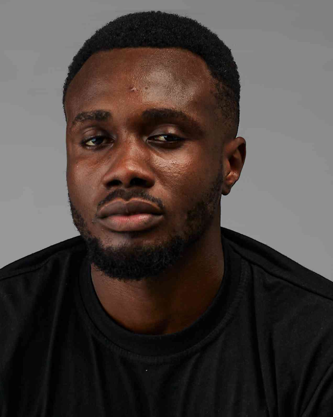 Close-up portrait of a young man with a serious, slightly skeptical expression. Plain grey background emphasizes facial features and mood.