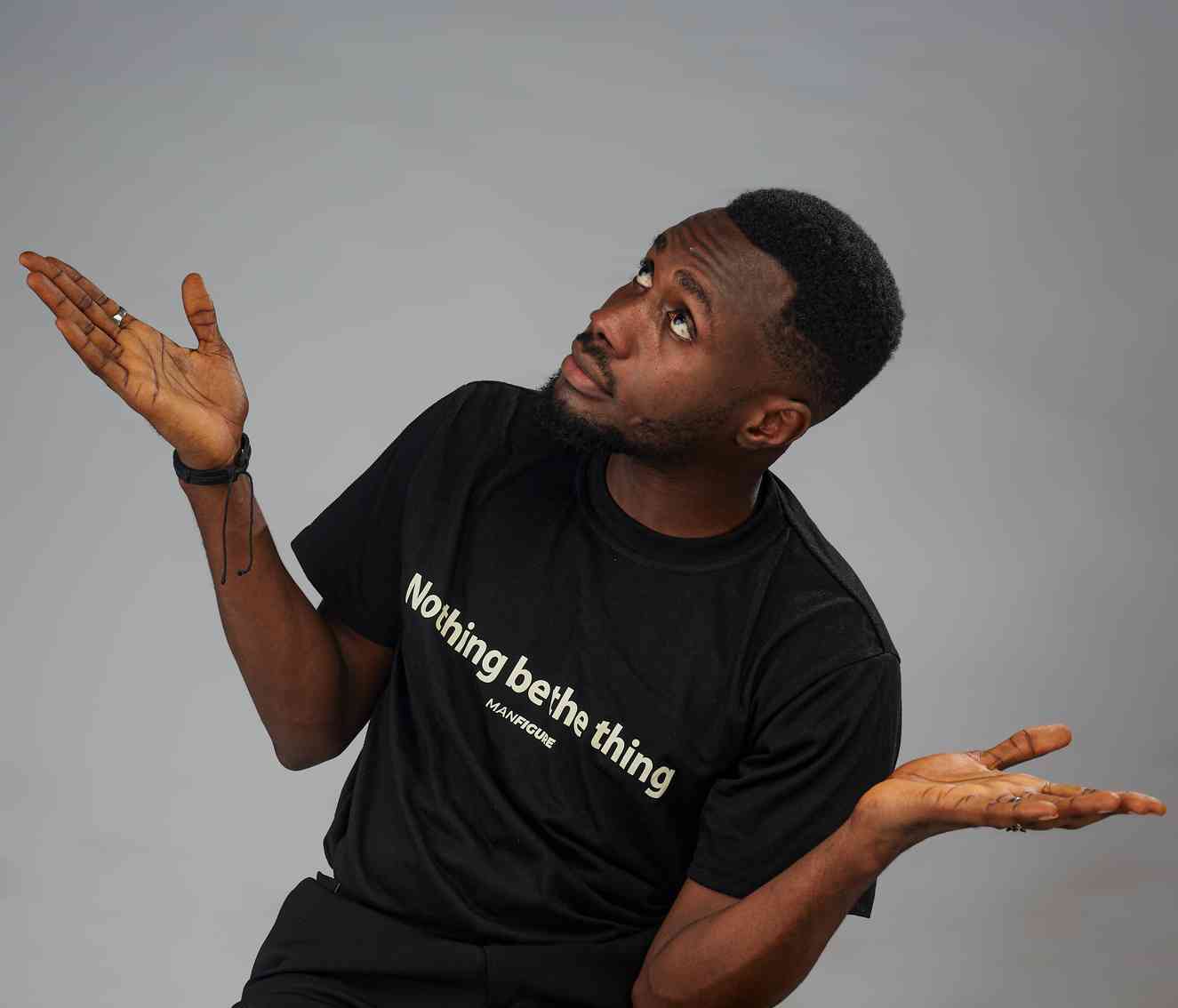 A young man in a black t-shirt shrugs his shoulders with palms up, looking upwards in a confused or uncertain expression. Neutral grey background.