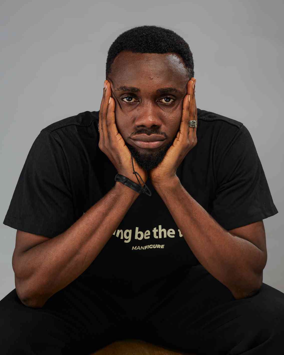 A man sitting with hands on his face, leaning forward and staring directly into the camera, showing a thoughtful or tired expression.