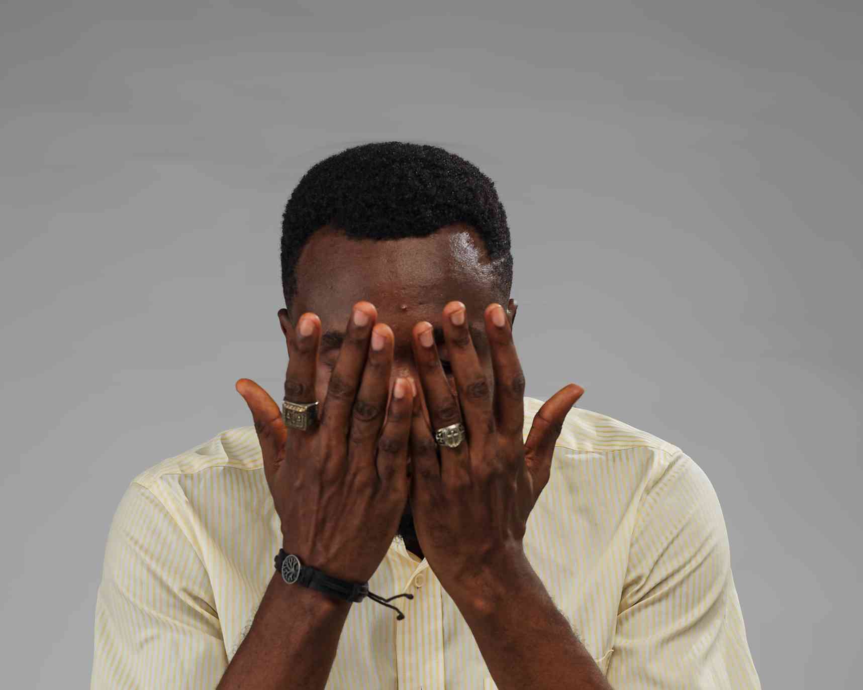 A man in a light yellow striped shirt covers his face with both hands, conveying emotions like shame, frustration, or sadness.