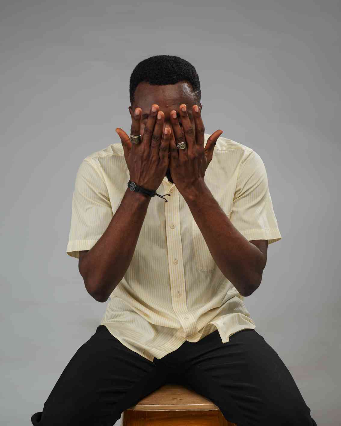 A man wearing a yellow striped shirt covers his face with both hands, expressing emotion or shyness in a seated position.