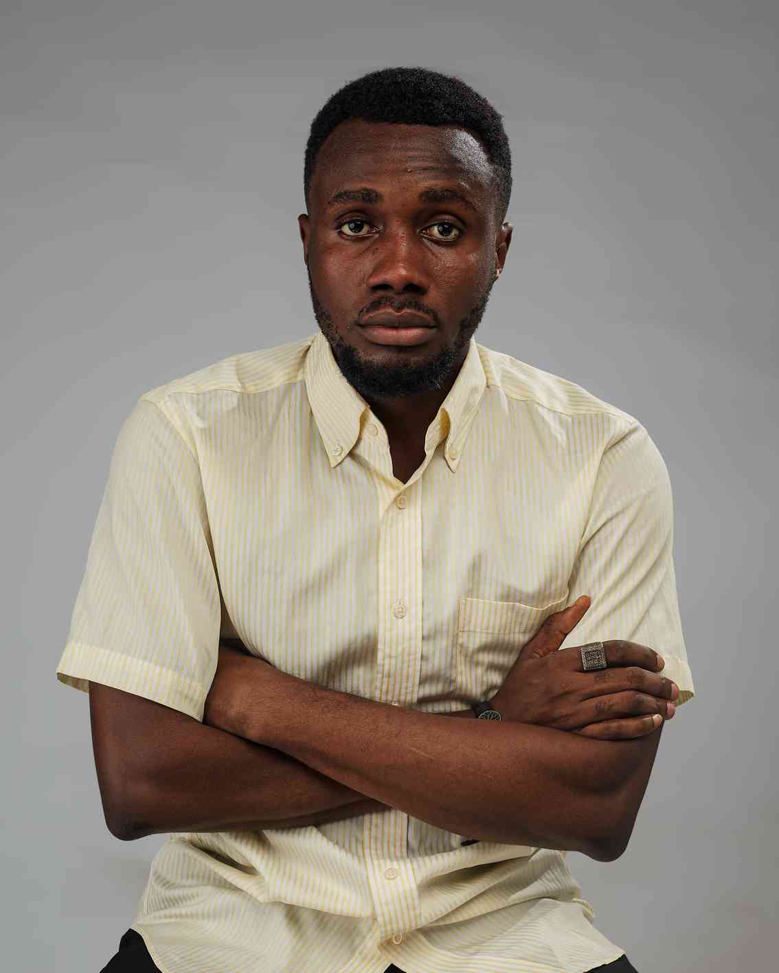 A serious-looking man with folded arms stares directly at the camera, wearing a striped short-sleeve shirt.