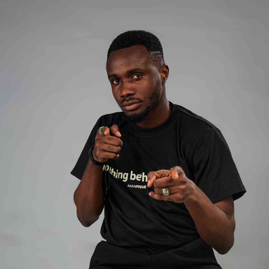 A confident man in a black motivational t-shirt poses with both hands pointing forward, smiling subtly.